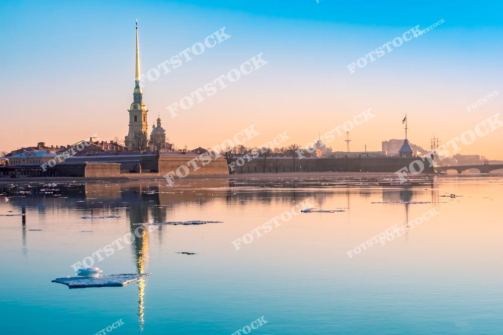 На фото — Петропавловская крепость, один из главных символов Санкт-Петербурга. Утренние лучи солнца освещают шпиль Петропавловского собора, отражающийся в зеркальной глади Невы. На реке дрейфуют отдельные льдины — предвестники наступающей весны. Вдали видны флаги, мачты и купола, создающие силуэт исторического центра города. Это место — отправная точка истории Петербурга и одна из его самых узнаваемых панорам.